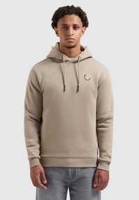 Jonge man in een beige hoodie met een klein, circulair tekstlogo op de borst, in combinatie met lichtgrijze jeans, staand tegen een effen witte achtergrond.