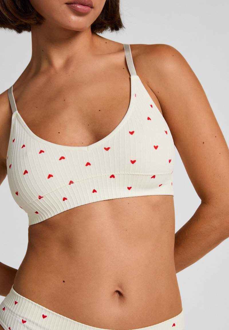 Licht crèmekleurig bralette met een geribbelde textuur, voorzien van kleine rode hartpatronen, dunne bandjes en een ronde halslijn.
