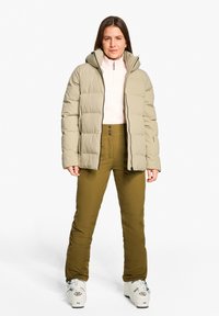 Giacca imbottita beige con cappuccio, sovrapposta a una maglietta rosa chiaro. Pantaloni verde oliva a vita alta con chiusura a bottoni. Stivali da sci bianchi.