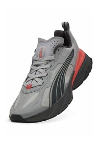 Chaussure de sport grise avec une tige en mesh, des accents noirs et un talon rouge. Forme arrondie avec des lacets proéminents et des détails texturés.