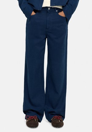 Osoba nosząca luźne, ciemnoniebieskie spodnie, brązowe mokasyny z czerwonymi sznurowadłami, ręce w kieszeniach oraz granatową kurtkę na białym swetrze.