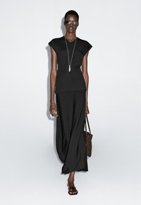 Große Frau, die ein schwarzes ärmelloses Oberteil, weit geschnittene Hosen, eine Sonnenbrille, eine Halskette, Sandalen trägt und eine braune Handtasche hält, geht vor weißem Hintergrund.