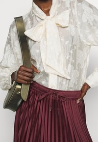 Blusa bianca trasparente con motivo floreale e fiocco, abbinata a una gonna plissettata bordeaux e una borsa verde oliva con accenti metallici.