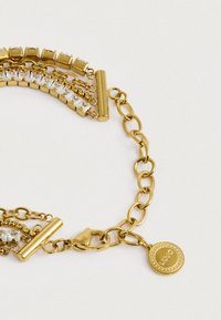 Βραχιόλι σε χρυσό τόνο με συνδεδεμένες αλυσίδες, καθαρές τετράγωνες πέτρες, κούμπωμα με αχίλλειο και στρογγυλό διακοσμητικό με λογότυπο σε λευκό φόντο.