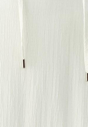 Acercamiento a una tela blanca texturizada con crestas verticales y dos cordones blancos que terminan en puntas de metal plateado.