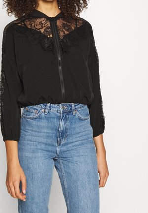 Zwarte rits-top met een kanten yoke en mouwen, gecombineerd met high-waisted blauwe jeans. De top heeft een ontspannen pasvorm en een gestructureerde stof.