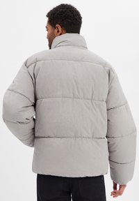 Graue Steppjacke mit hohem Kragen und voluminöser Silhouette. Weicher, strukturierter Stoff mit sichtbaren Nähten und großen Polsterabschnitten.