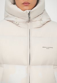 Krämfärgad pufferjacka med hög krage, dragkedja fram och logotypdetalj. Har elastiska snoddar och kviltad textur.