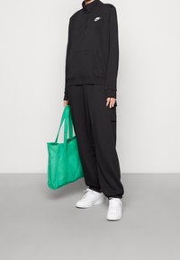 Svart zip-up hoodie med framficka och Nike-logotyp, kombinerad med matchande svarta byxor. Håller en ljusgrön tote-väska och har på sig vita sneakers.