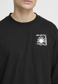 Black long-sleeve t-shirt made of cotton. Features white graphic with sun and waves, text "UNLIMITED SUMMER" on left chest area. Regular fit.