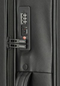 Valigia nera con serratura a combinazione con logo TSA, superficie testurizzata, doppie cerniere e marchio "GUESS" sugli estrattori delle cerniere.
