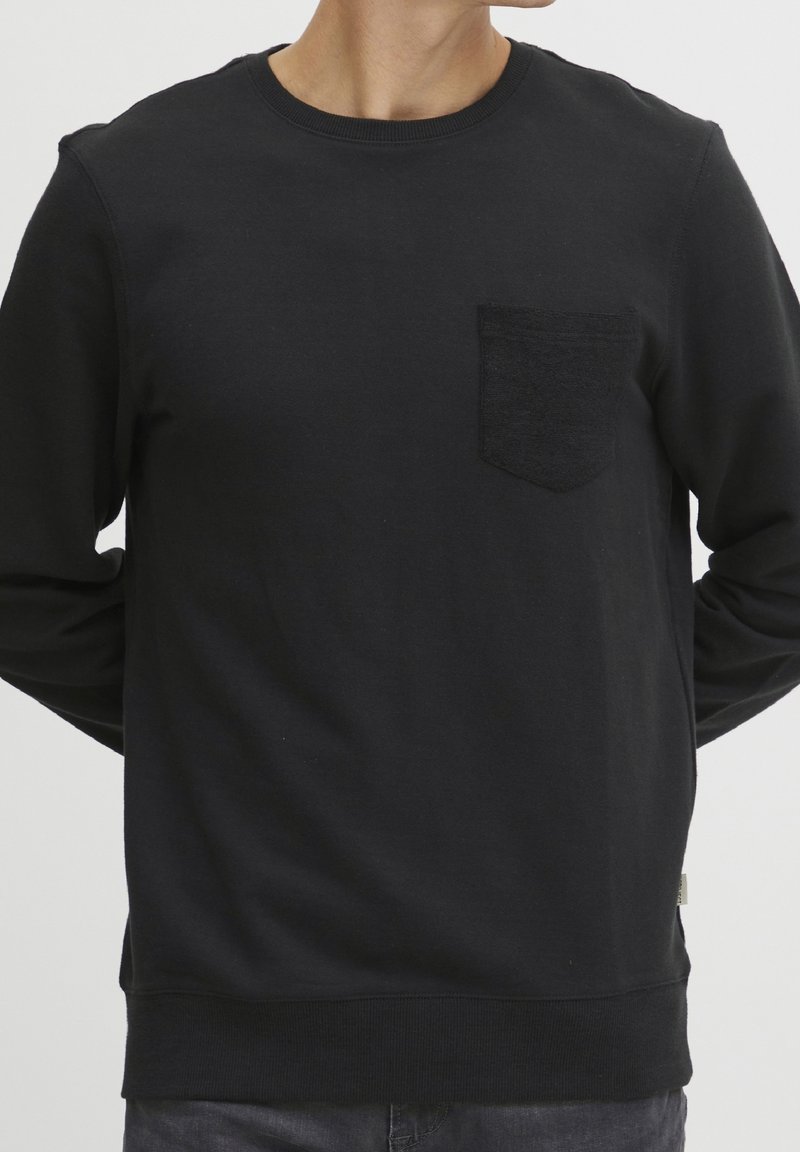 Adulte portant un t-shirt noir à manches longues et col rond, avec une poche poitrine, debout les mains derrière le dos.