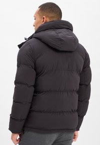 Zwarte pufferjack met een capuchon, uitgevoerd met horizontale stiksels en een gladde textuur. Bevat een logo-opdruk op de linkerschouder.