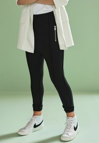 Pantaloni negri strâmți cu buzunar lateral cu fermoar, asortați cu adidași albi cu guler înalt, având un swoosh negru, pe un fundal verde.