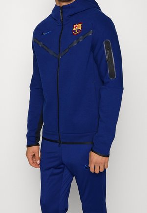 Mężczyzna ubrany w niebieską kurtkę na zamek i pasujące spodnie z logo FC Barcelona i Nike, stojący na jednolitym białym tle.