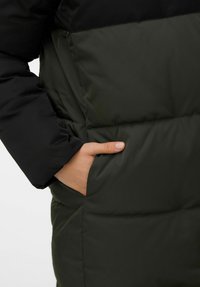 Paminkštinta striukė su juoda viršutine dalimi ir tamsiai žalia apatine dalimi. Yra šoninė kišenė ir lygi, blizgi tekstūra.