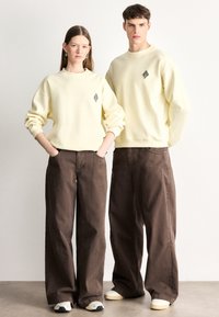 Kremas spalvos džemperis su juodu geometriniu logotipu, derinamas su laisvais rudais kelnėmis. Audinys atrodo minkštas, su laisvu kirpimu ir plačiu kojų atvartų atidarymu.