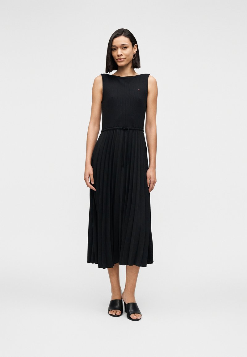 Femme debout portant une robe midi plissée noire sans manches avec une ceinture à la taille et des sandales noires à enfiler sur un fond clair uni.