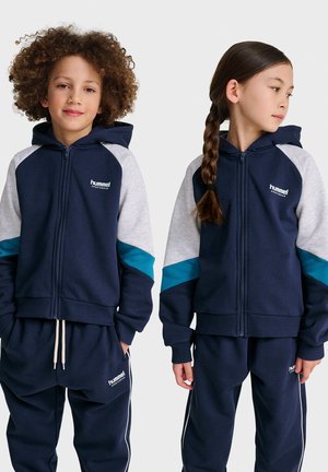 Marineblauwe zip-up hoodie met lichtgrijze mouwen en turquoise accenten, gecombineerd met marineblauwe joggingsbroek. Bevat logodetails en heeft een casual pasvorm.