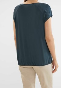 Tmavě modrý top s krátkým rukávem, volného střihu, hladkým povrchem, zaobleným výstřihem a nařaseným lemem, který je kombinován se světle béžovými kalhotami.