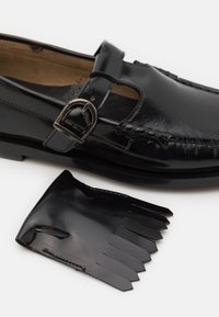 Schwarzer Lederschuh mit einem Riemen und einer Metallschnalle, mit abgerundetem Zehenbereich und genähten Details. Begleitet von einem passenden Fransenaccessoire.