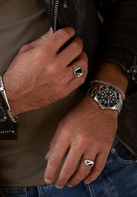 Montre en argent avec cadran noir, chaîne et bague. Le design présente des accents géométriques, des détails texturés et des matériaux contrastants. Veste en cuir visible.
