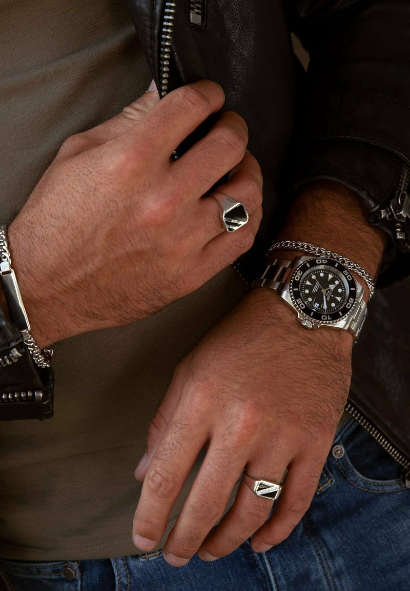 Montre en argent avec cadran noir, chaîne et bague. Le design présente des accents géométriques, des détails texturés et des matériaux contrastants. Veste en cuir visible.