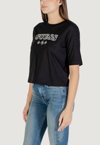 T-shirt nera cropped a maniche corte, con il logo "GUESS" in evidenza in un font metallico e decorazioni floreali.