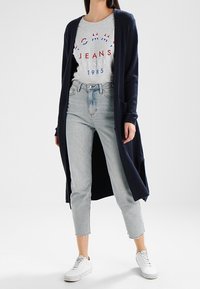 Osoba ubrana w granatowy długi kardigan, szary t-shirt z logo Tommy Jeans, jasnoniebieskie cięte dżinsy i białe trampki na białym tle.