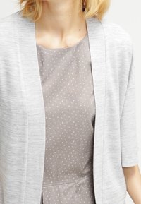 Cardigan grigio chiaro indossato sopra un vestito grigio a fantasia con piccoli dettagli bianchi. Il cardigan presenta una vestibilità rilassata e un design aperto davanti.