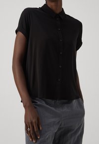 Black short-sleeve, collared shirt with a button-down front, made from lightweight fabric. Features rolled sleeves and a relaxed fit.