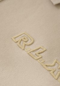 Beige stoffen oppervlak met een opvallend, geborduurd logo in lichtgele draad, dat ingewikkelde stiksels en een gestructureerde weving laat zien.