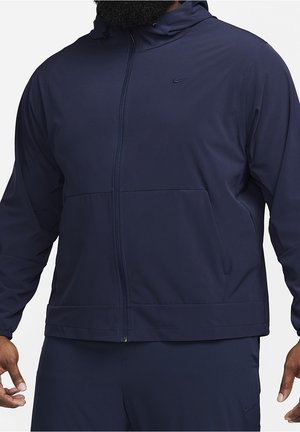 Homme portant une veste à capuche zippée Nike bleu foncé et un pantalon assorti sur fond uni.