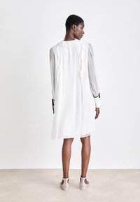 Weißes Kleid mit Rüschendetails, geflochtenem Rücken und langen, halbtransparenten Ärmeln. Begleitet von hellen Schuhen mit Ausschnitten.