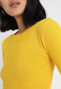 Yellow ribbed long-sleeve top with a round neckline. The textured fabric creates a subtle pattern. The fit is snug, highlighting the body shape.