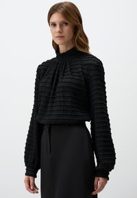 Zwarte blouse met textuur en hoge hals, voorzien van geplooide stof en lange mouwen, gecombineerd met een strakke zwarte rok.