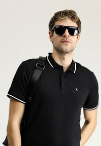 Polo nero con bordini bianchi col colletto e maniche, con un piccolo logo sul petto. Il modello porta una borsa a tracolla nera.