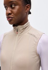 Γυναίκα που φοράει μπεζ αμάνικο γιλέκο με φερμουάρ και το λογότυπο «CASTELLI» στον ώμο, πάνω από λευκό μακρυμάνικο μπλουζάκι, κοιτάζοντας προς τα αριστερά.