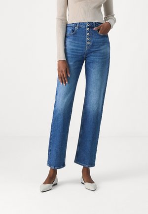 High-waisted, straight-leg blue denim jeans with a light wash, five-button fly, and side pockets; paired with a beige ribbed sweater and white shoes.