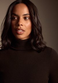 Primer plano de una persona con un jersey de cuello alto de color marrón oscuro, de tejido acanalado. Tiene el cabello ondulado y a la altura de los hombros, piel clara y maquillaje sutil.