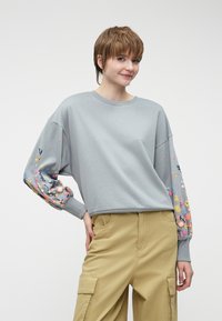 Pelēka sporta krekla ar krāsainiem ziedu izšuvumiem uz piedurknēm, maiga tekstūra, brīvi pieguļoša forma, apaļš kakls. Apvienots ar khaki krāsas militārā stila biksēm.