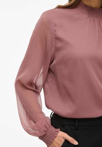 Blusa de color rosa en tejido transparente con mangas abullonadas y puños fruncidos, que presenta un cuello alto y un acabado texturizado.