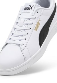 Tenis sintético branco com uma faixa lateral de pele preta, detalhes do logótipo em dourado e uma sola de borracha texturizada. Apresenta um sistema de atacadores tradicional.