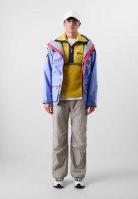 Blauwe waterdichte jas met roze accenten, gele fleece eronder, en grijze broek. Zwart-witte sneakers maken de outfit compleet.