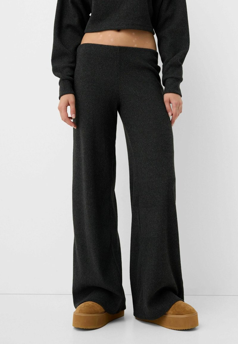 Personne portant un pantalon noir côtelé à jambes larges, un pull noir court et des chaussures à plateforme marron, debout sur un fond blanc.