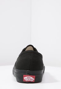 Baskets en toile noires à bout rond, avec étiquette logo rouge contrastante au talon, semelle plate noire et détails de couture visibles sur la tige.