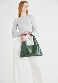 Green leather handbag with a rectangular cutout handle, featuring a smooth texture and minimalist design, held by a model in white pants.