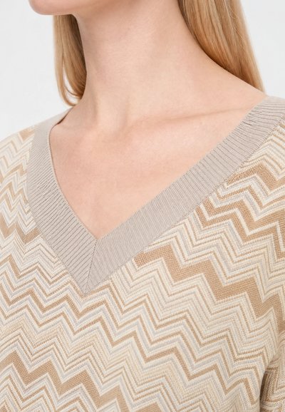 Gros plan d'une personne portant un pull à col en V beige et blanc avec un motif en zigzag, avec de longs cheveux blonds visibles.