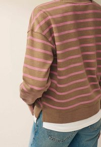 Felpa marrone con strisce rosa, caratterizzata da una vestibilità rilassata, polsini a costine e un orlo curvo. Indossata sopra una camicia bianca e jeans in denim.