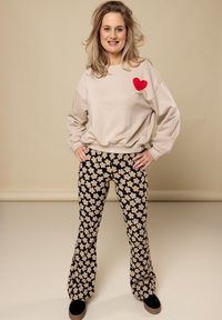 Beige sweatshirt met een rode hartgrafiek, gecombineerd met zwarte flare-pants met een wit bloemenpatroon. Model draagt zwarte sneakers.